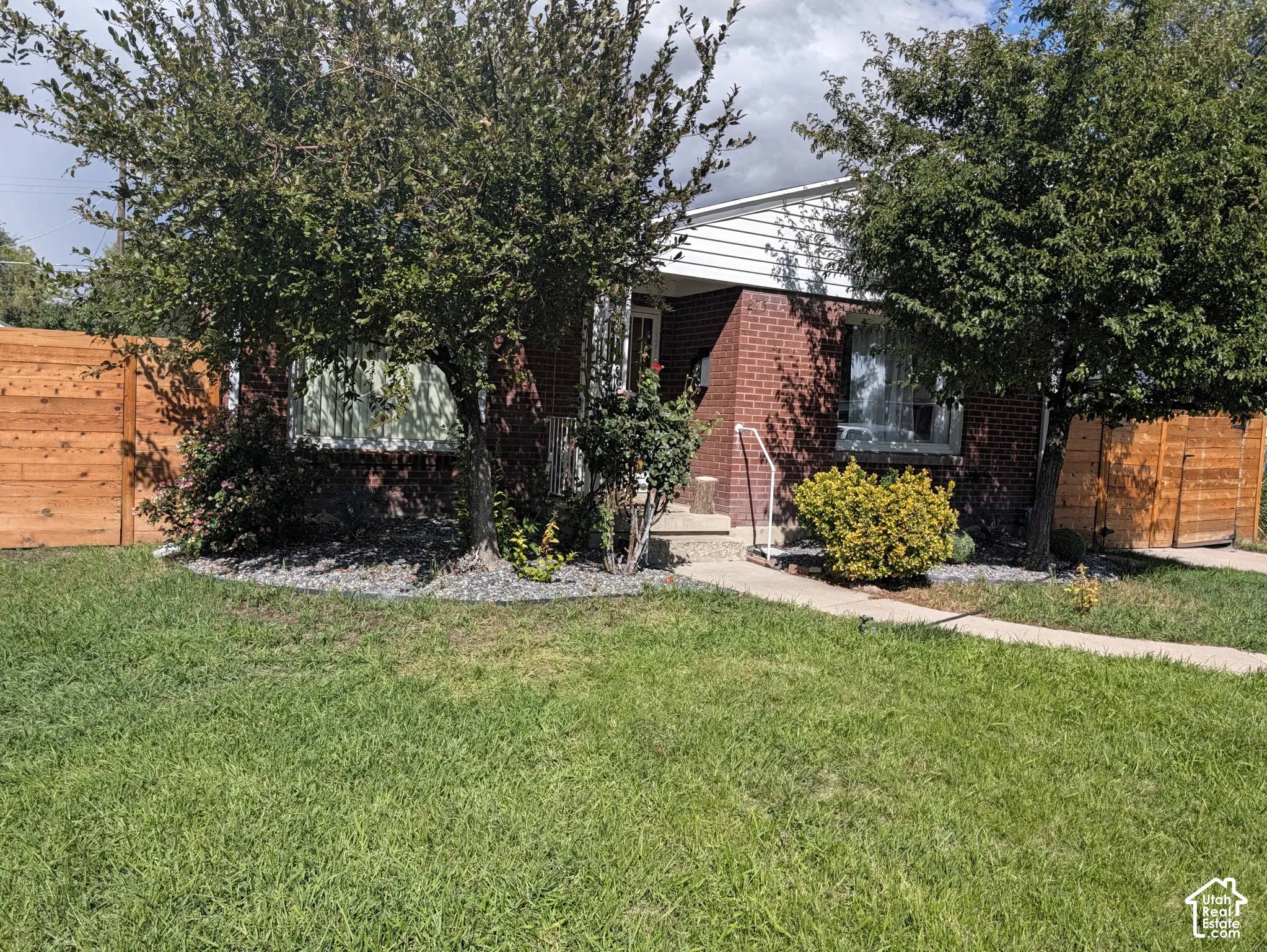 Brick exterior with white siding accents, mature trees, wood privacy fencing, and concrete walkway