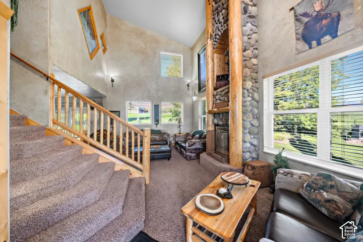 Vaulted ceiling with textured walls, stone-clad fireplace with wood accents, multiple windows with blinds, and a carpeted staircase with a wood railing