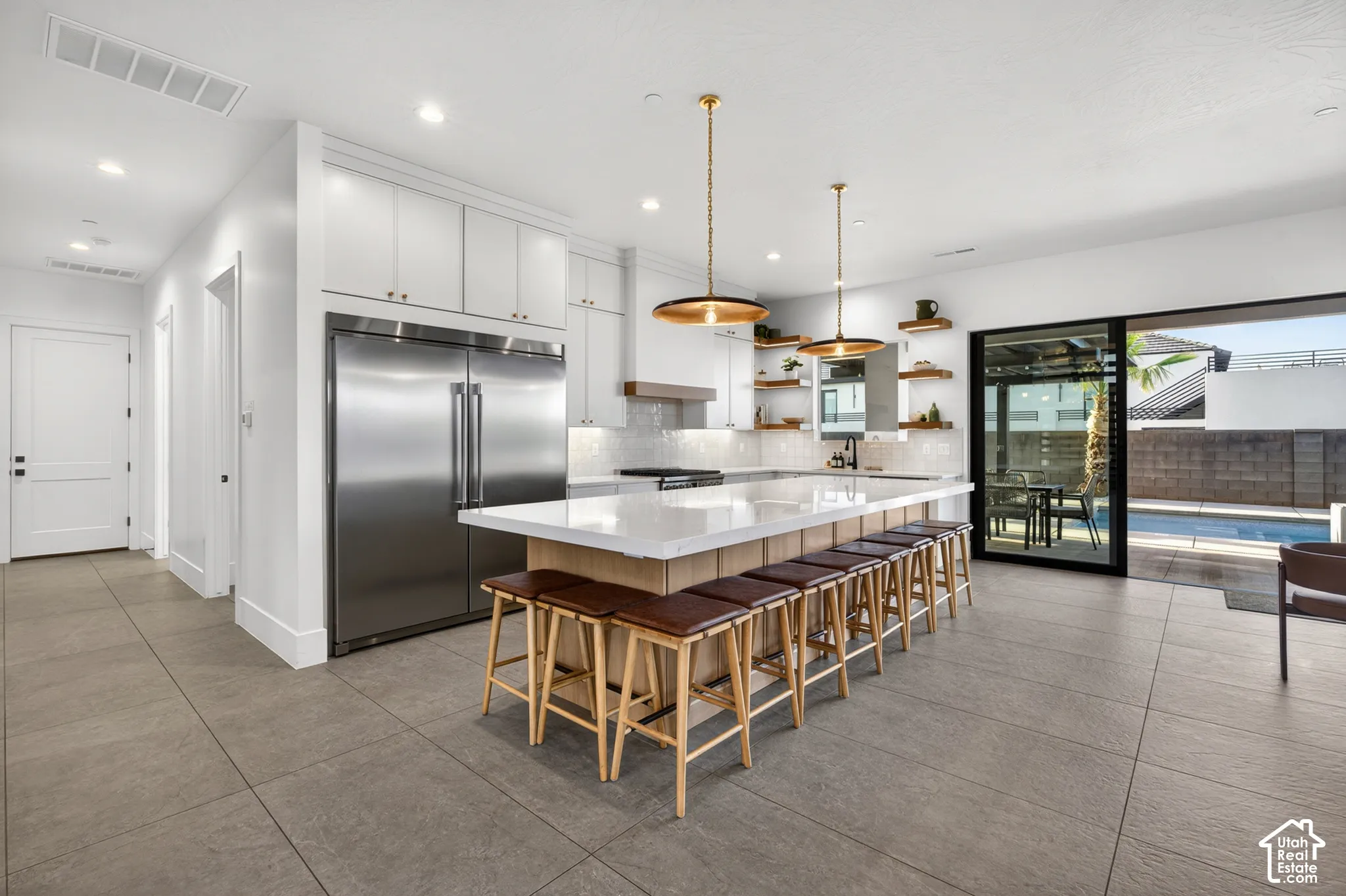 White cabinetry with brass hardware, stainless steel refrigerator, large kitchen island with counter seating, pendant lighting, and a sliding glass door to an outdoor pool
