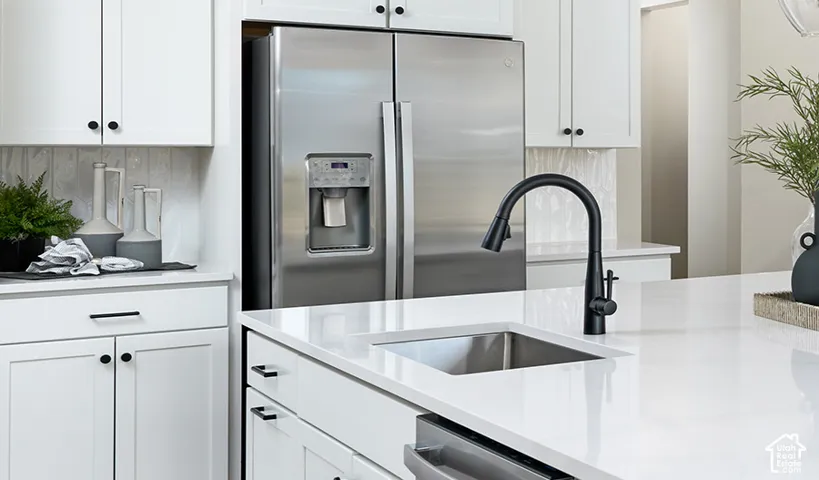 Stainless steel side-by-side refrigerator, white cabinetry with dark hardware, light countertop surfaces, stainless steel undermount sink, and a matte black pull-down faucet
