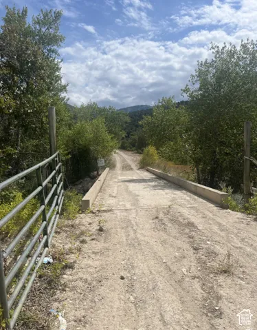 Gated dirt road with a concrete bridge, surrounded by mature trees and distant hills under a cloudy sky