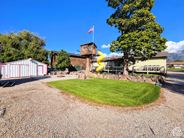Spacious gravel area, manicured lawn, white and red utility building, mature tree, and a treehouse structure with a yellow slide