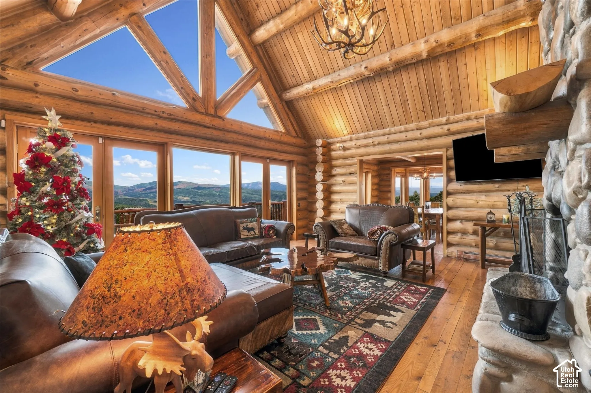 Expansive great room featuring a stone fireplace, wood-finish flooring, exposed log walls, and a vaulted ceiling with triangular window elements