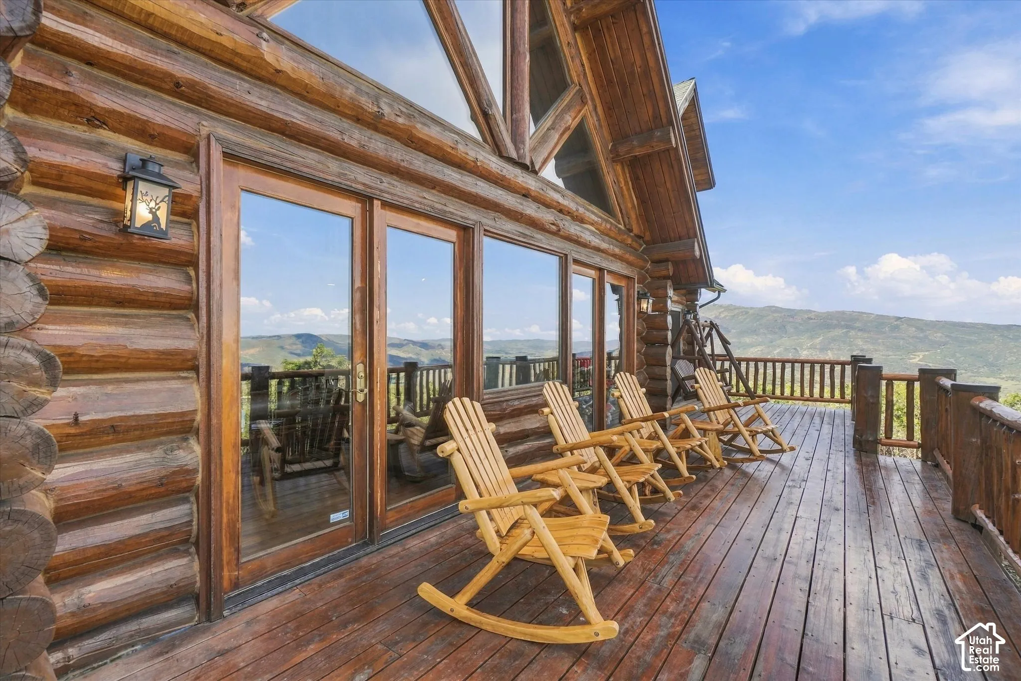 Expansive wood deck with a full wood railing, featuring a log cabin exterior, large glass doors, and a decorative outdoor wall light