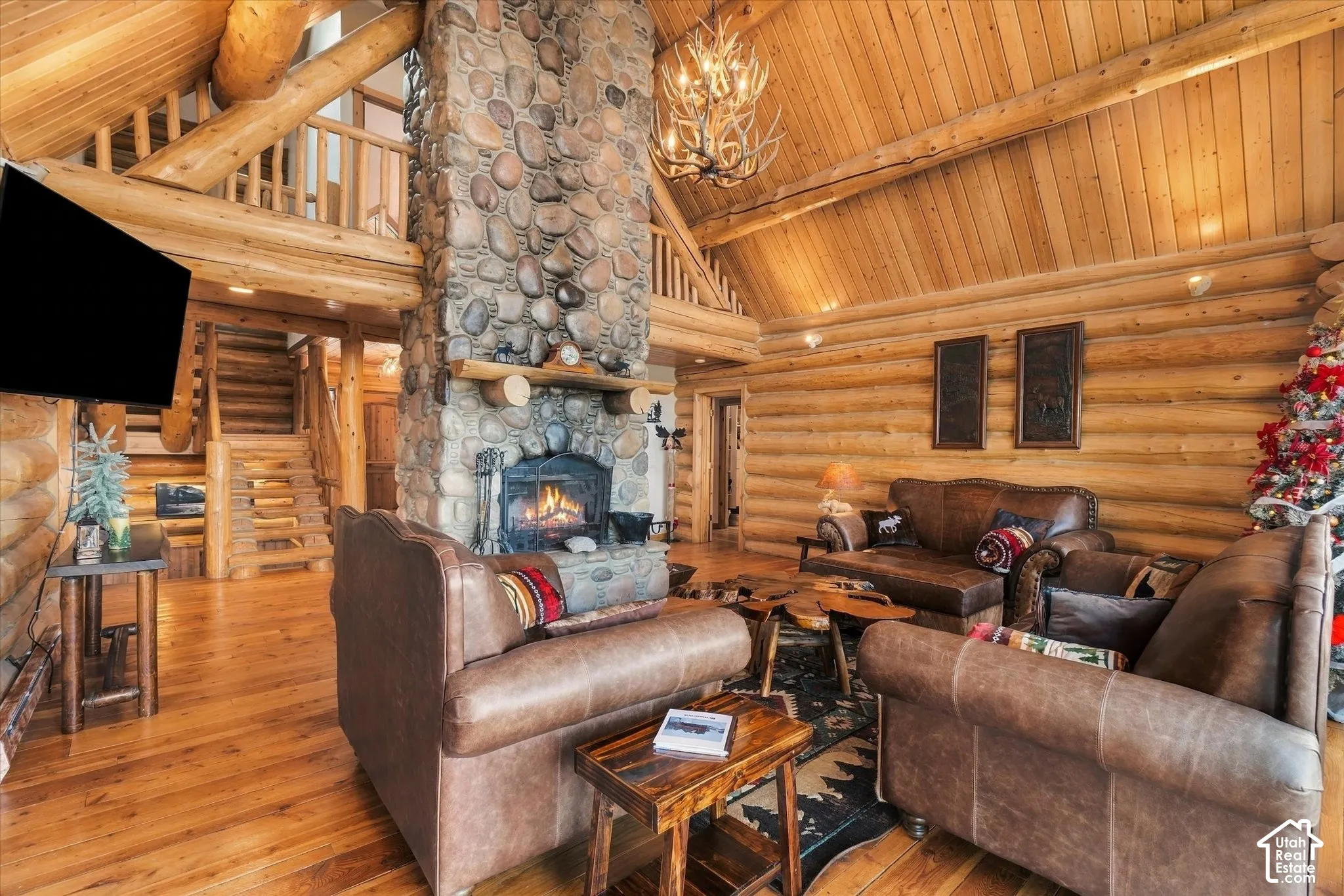 Living area featuring a stone-clad fireplace, log walls, wood-finish flooring, a vaulted wood-paneled ceiling, and a built-in staircase