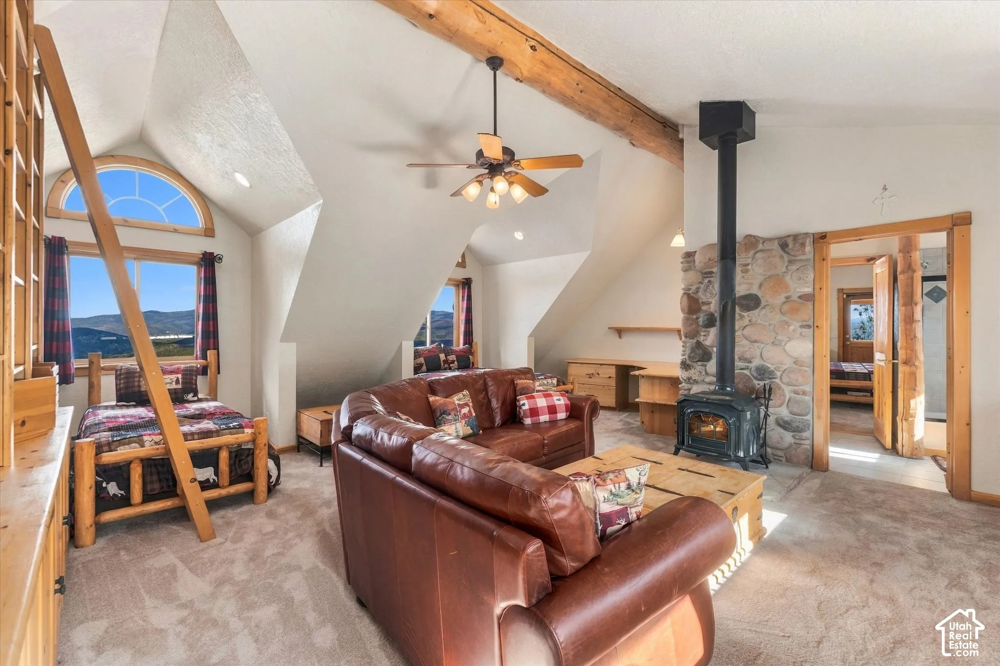 Vaulted ceiling with exposed wood beam, stone-clad wood-burning stove, dormer windows, ceiling fan, and light-toned carpeting