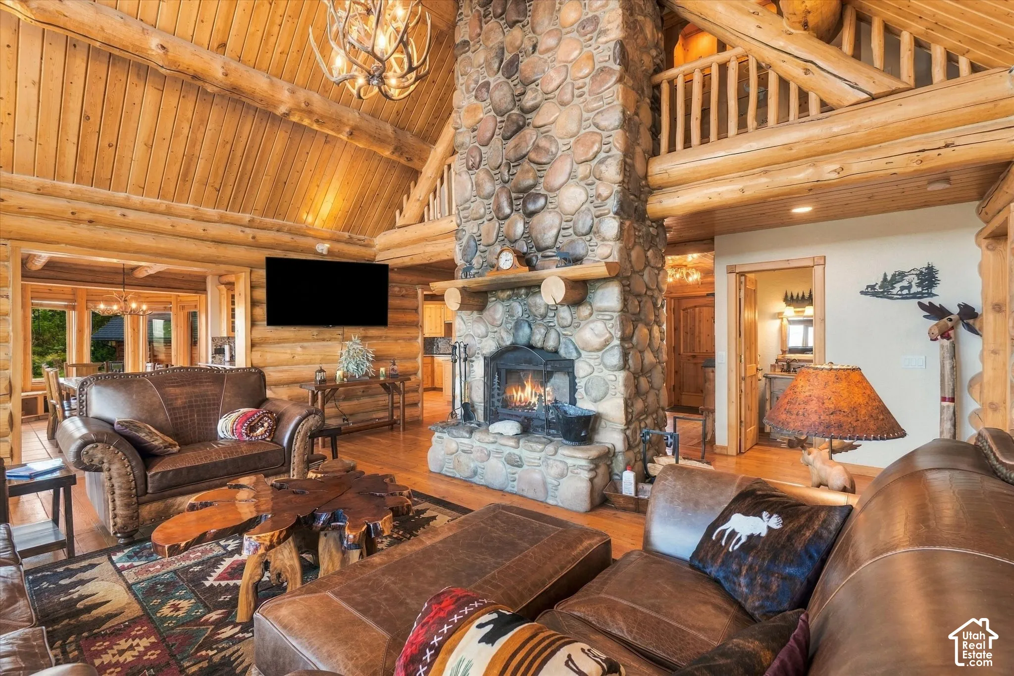 Vaulted wood-paneled ceiling, exposed log beams, floor-to-ceiling stone fireplace, wood-finish flooring, and a log-style balustrade