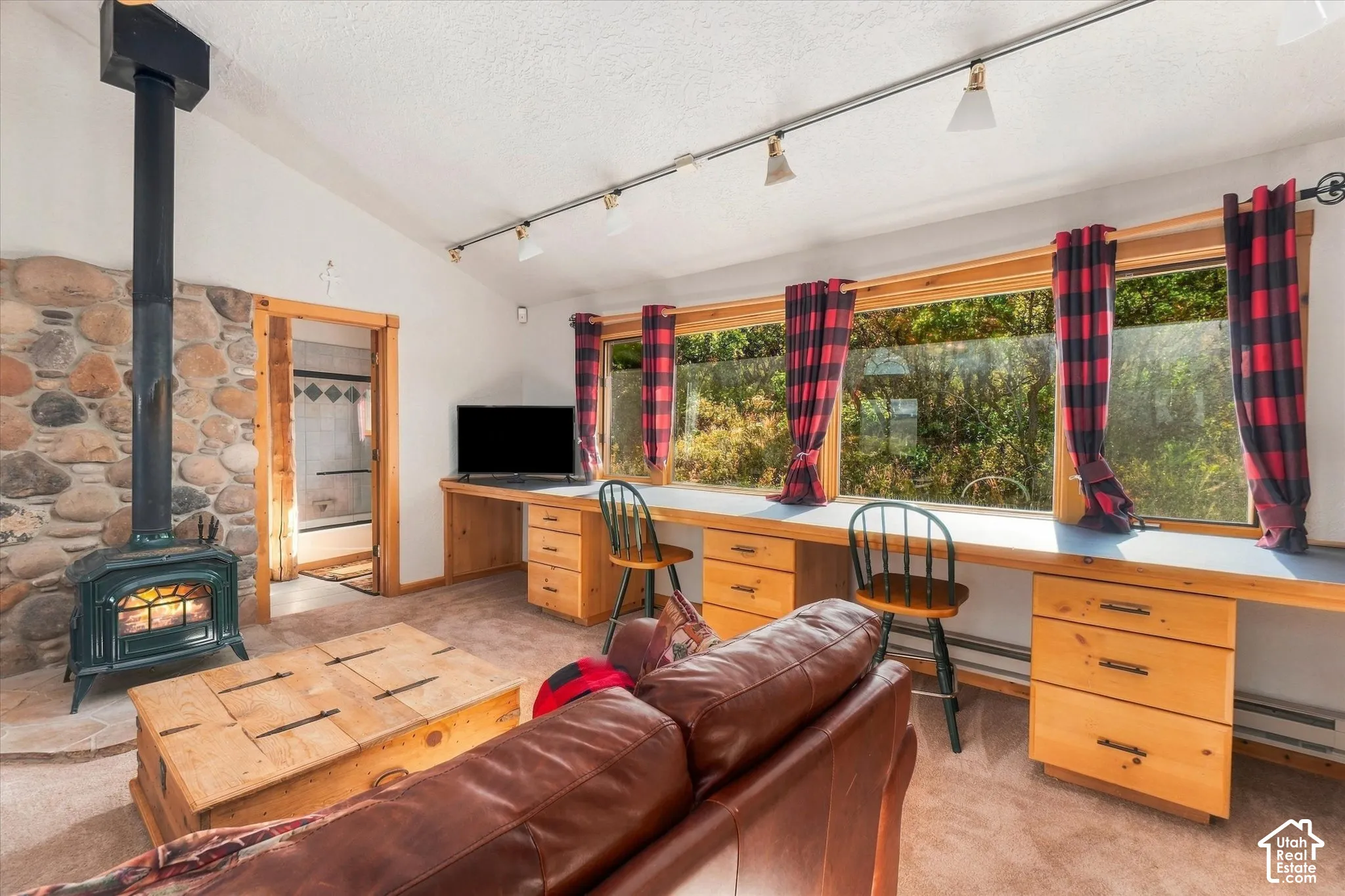Vaulted ceiling, stone accent wall, wood-burning stove, track lighting, and built-in desk cabinetry