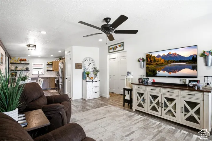 Recessed lighting, a ceiling fan with integrated lighting, wood-finish flooring, a kitchen area with stainless steel appliances, and a built-in entertainment console