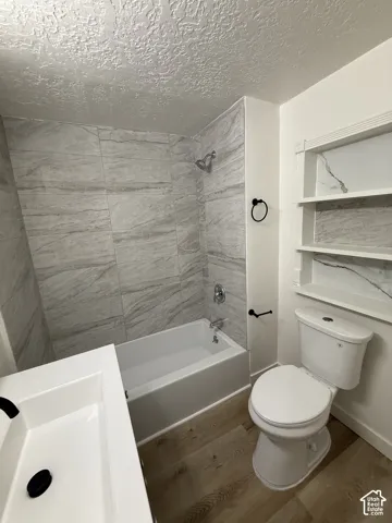 Full bathtub with a polished chrome shower head, wood-finish flooring, built-in shelving with stone-look backing, and a modern vanity with integrated sink