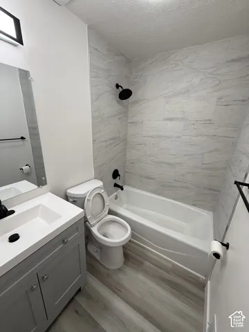 Bathroom featuring a white bathtub, tiled shower surround, gray vanity with integrated sink, black fixtures, and wood-finish flooring
