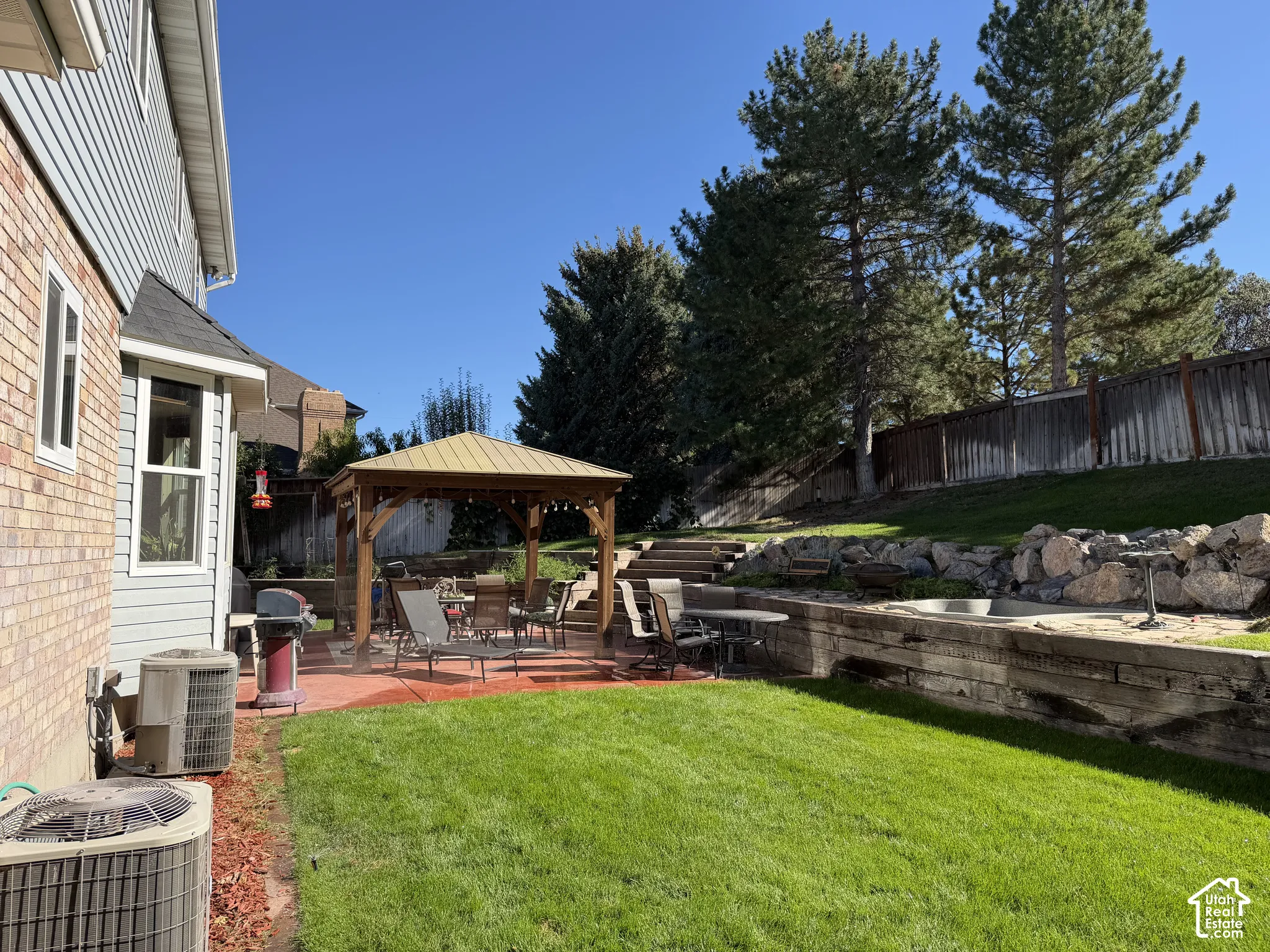Expansive backyard featuring a manicured lawn, a paved patio with a built-in gazebo, and terraced landscaping with mature trees and a wood retaining wall