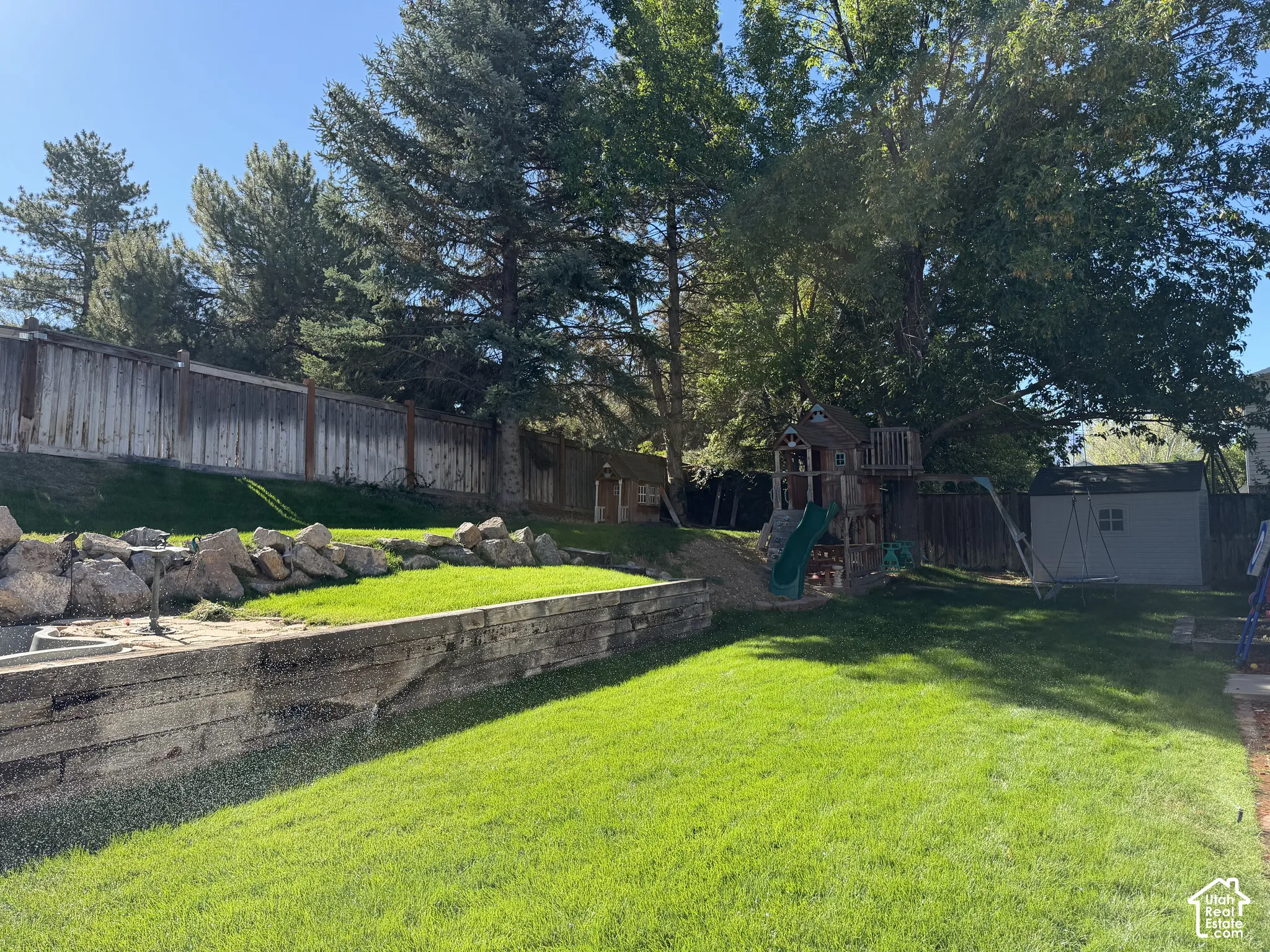 Expansive backyard featuring a tiered lawn, mature trees, wood retaining walls, and a wood privacy fence