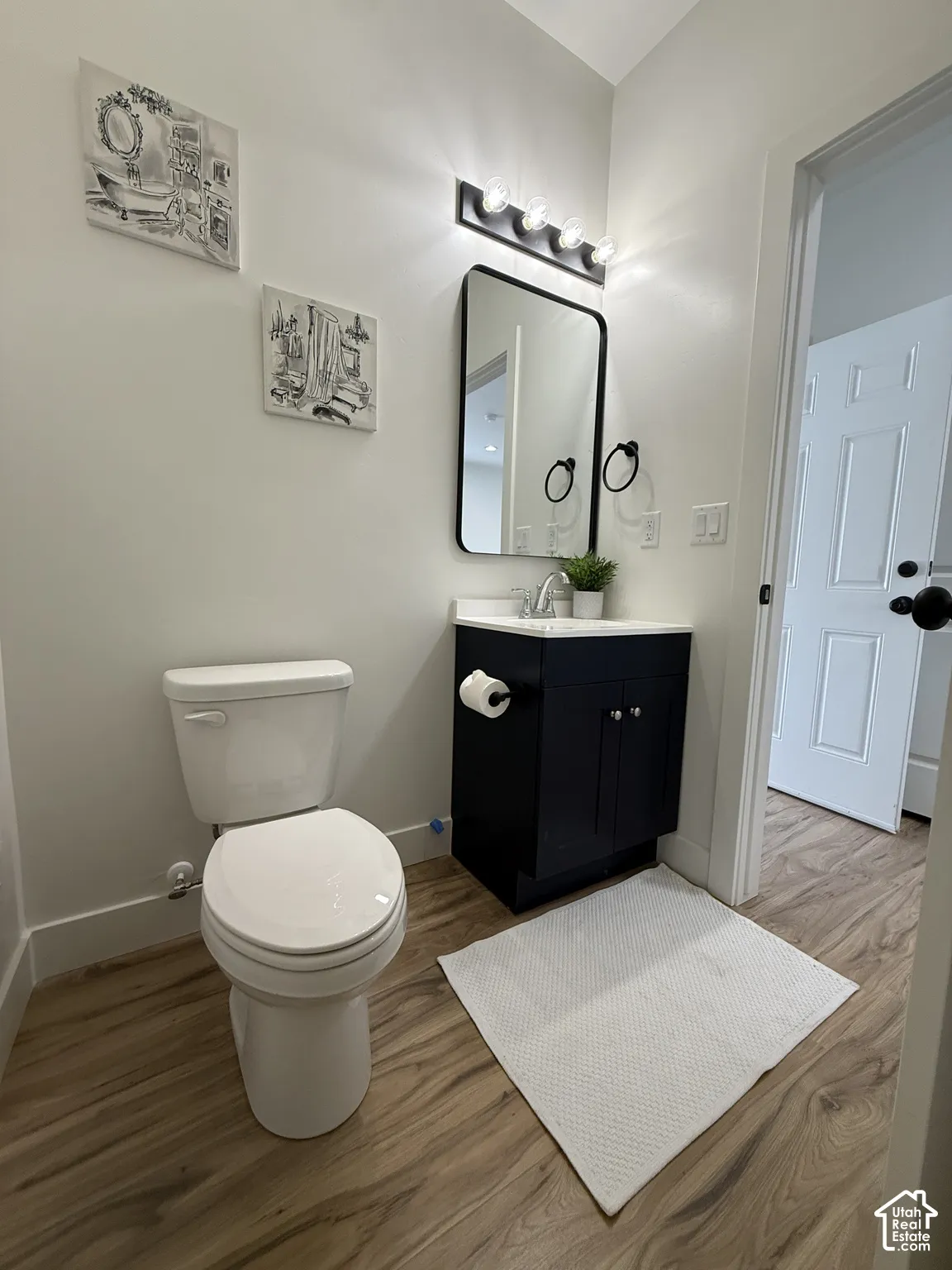 Wood-finish flooring, white toilet, dark vanity with white countertop, rectangular mirror with black frame, and a wall-mounted light fixture with four bulbs