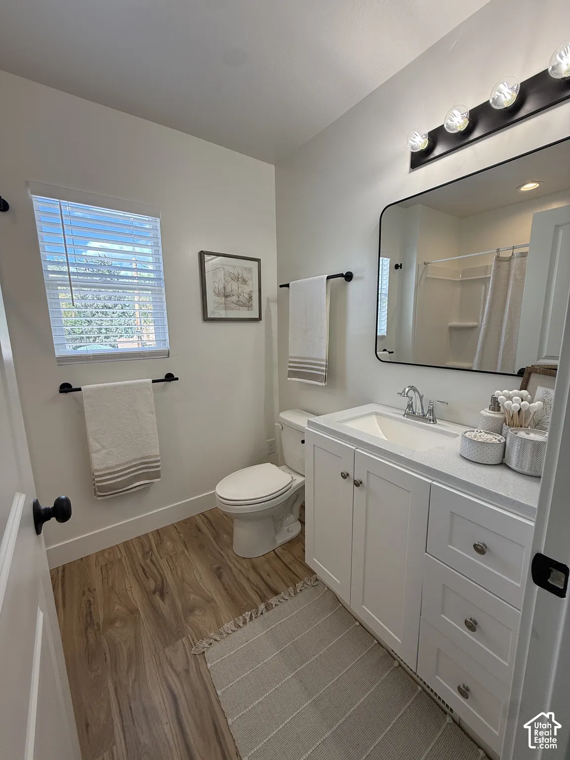 Wood-finish flooring, white vanity with integrated sink, black framed mirror, modern black light fixture, and a window with blinds