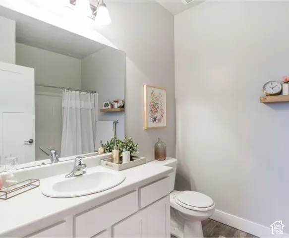 White vanity with an integrated sink and chrome faucet, full-width mirror, toilet, and wood-finish flooring