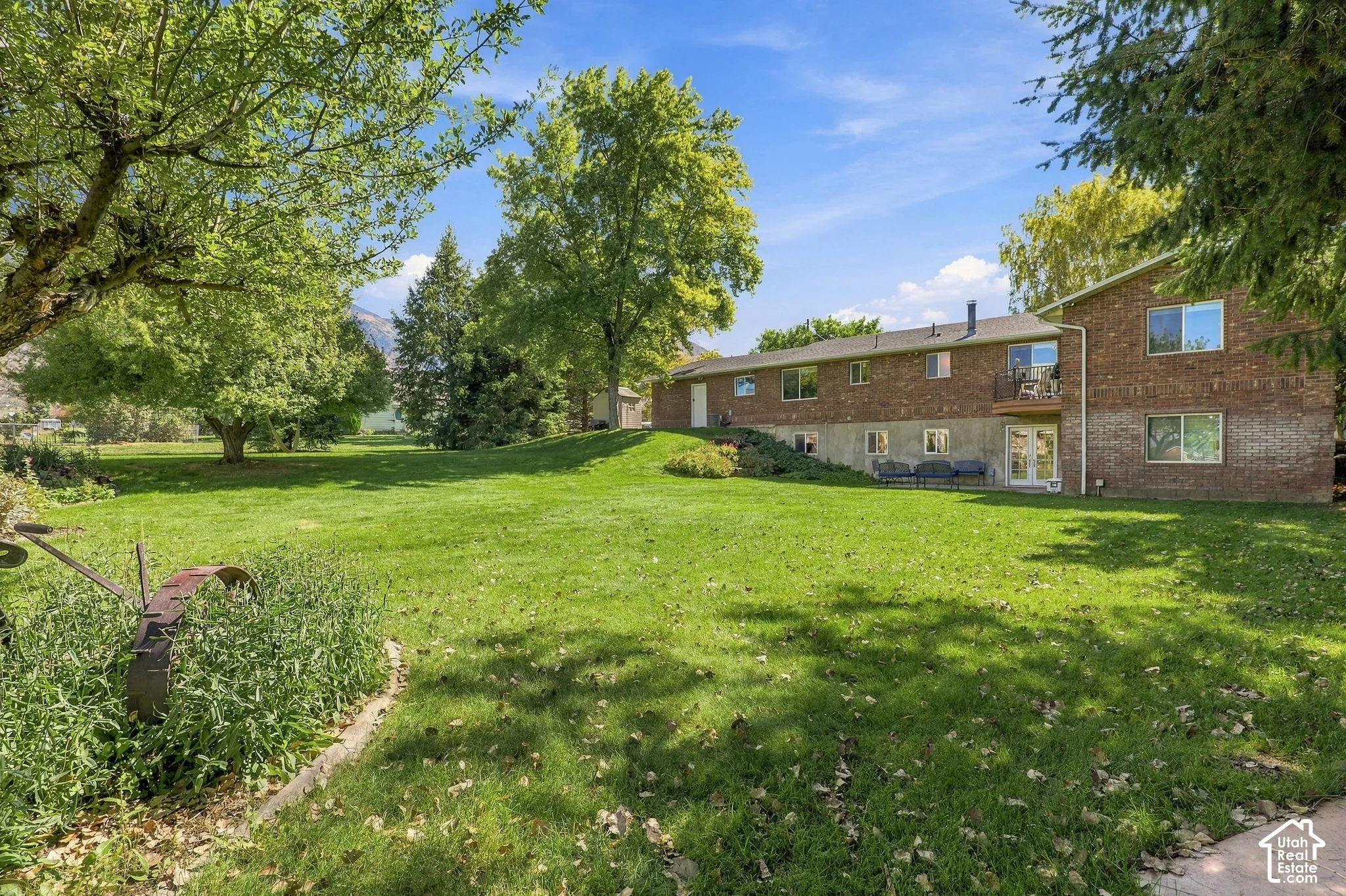 Expansive landscaped lawn, mature trees, brick exterior, upper-level balcony, and lower-level patio access