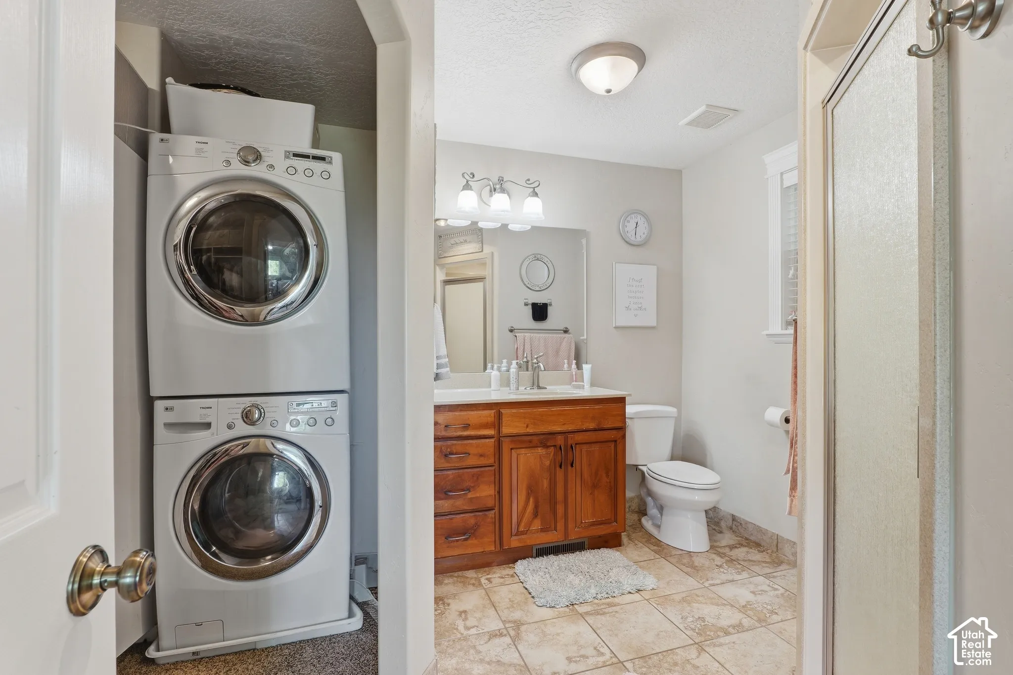 Integrated stacked laundry appliances, wood-finish vanity with countertop, frameless mirror, tiled flooring, and a shower enclosure