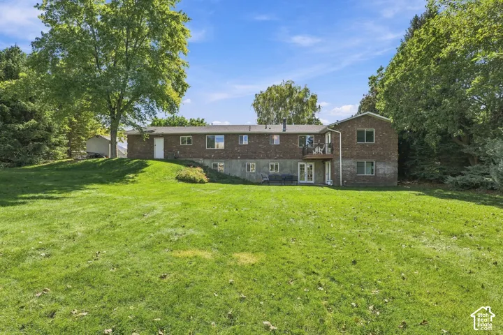 Expansive green lawn with mature perimeter trees, brick exterior, upper-level balcony, and lower-level patio doors