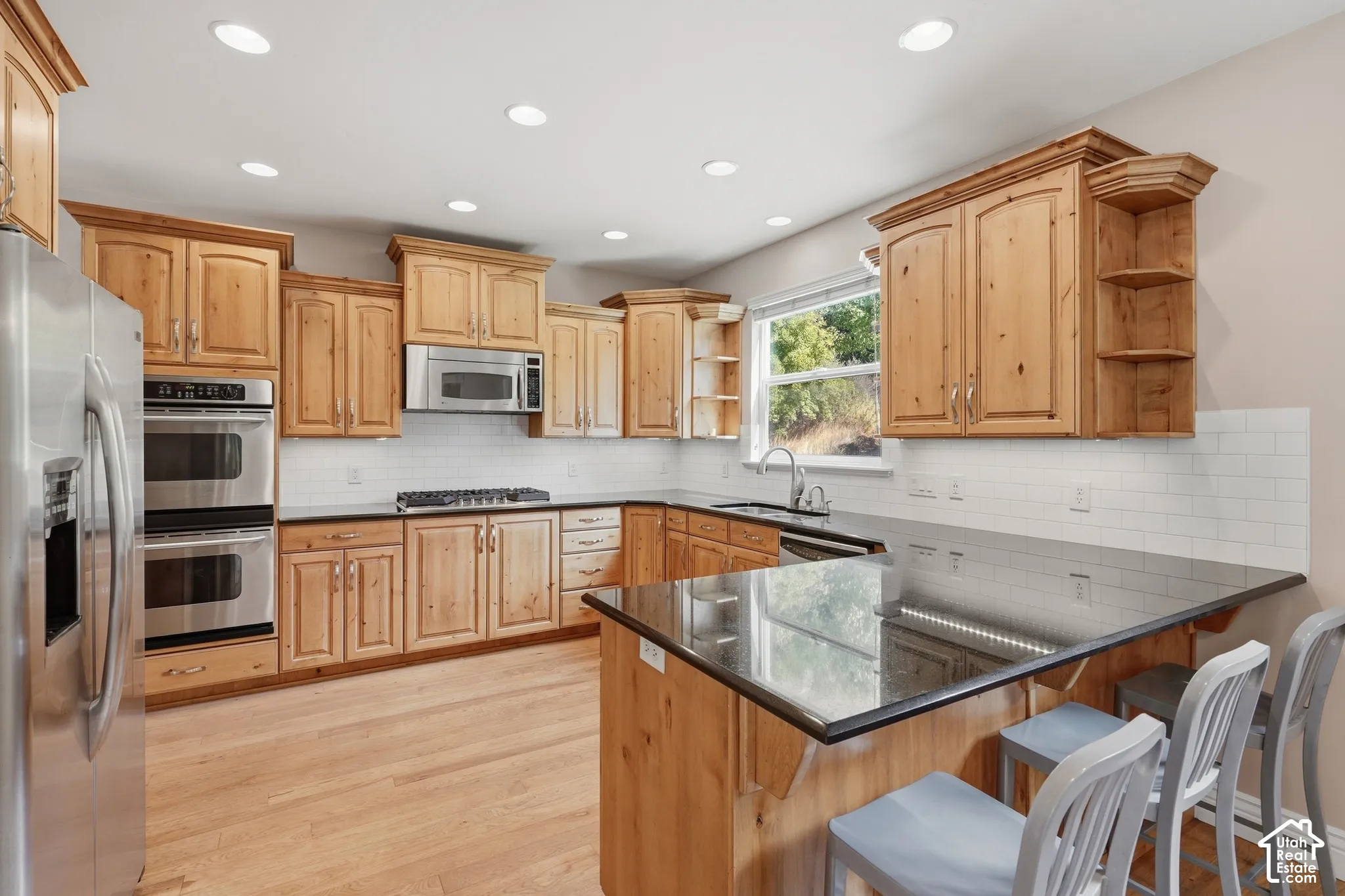 Kitchen featuring wood-finish cabinetry, stainless steel appliances, black countertops, white subway tile backsplash, and light wood-finish flooring