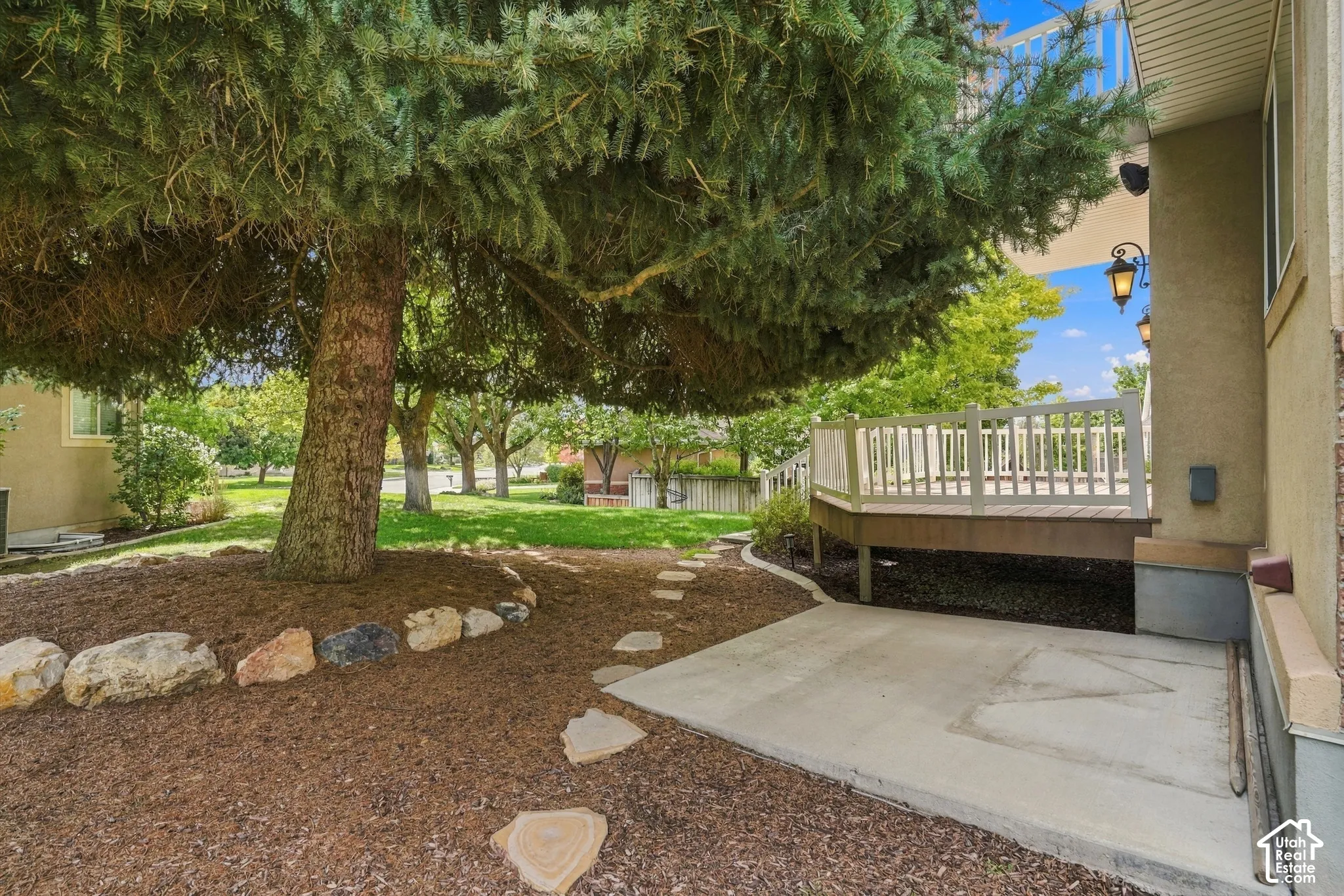Raised wood deck with white railing, concrete patio, mature tree with wood chip landscaping, stone pathway, and exterior wall sconces