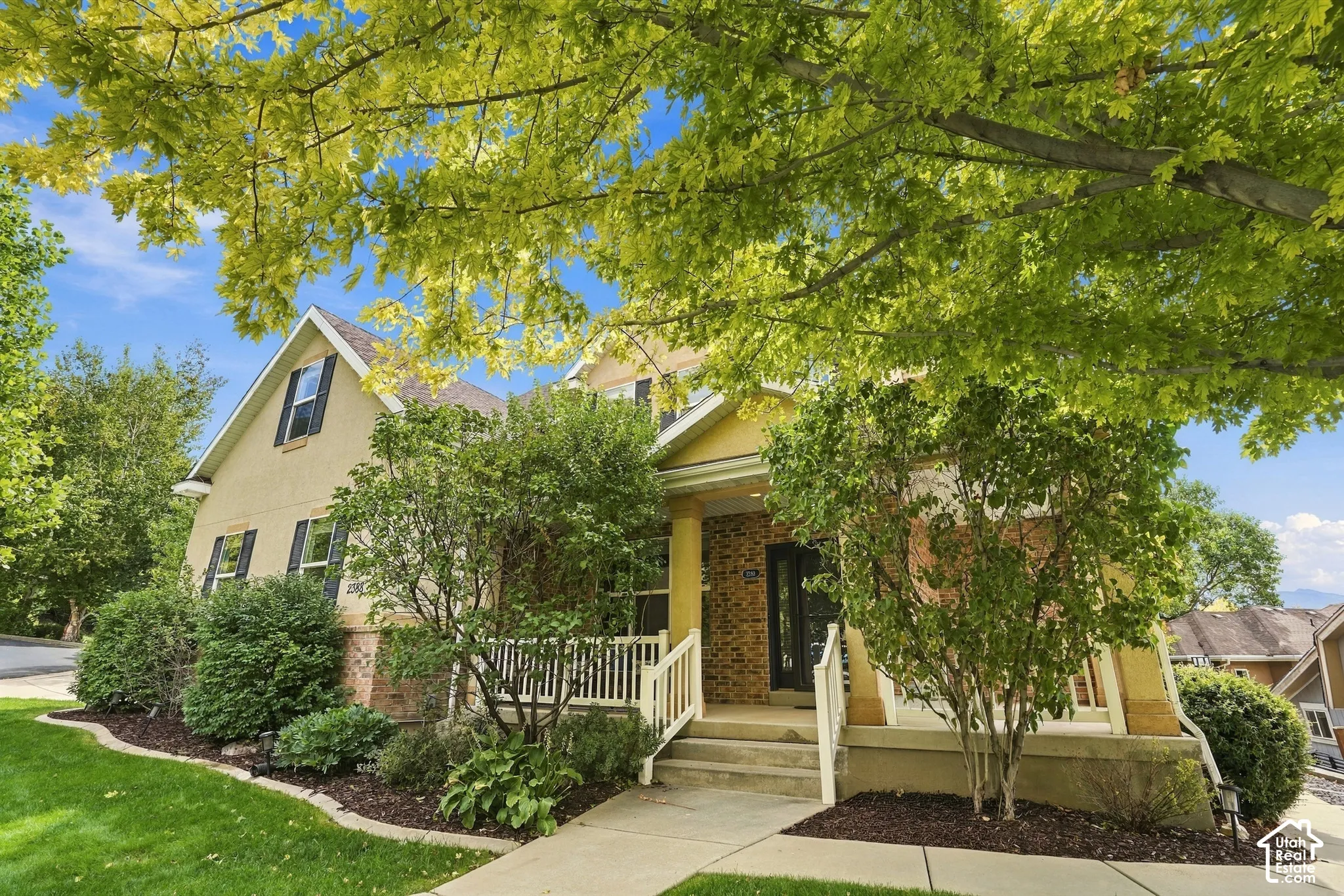 Stucco and brick exterior with a prominent front entrance, white railings, mature trees, and established landscaping