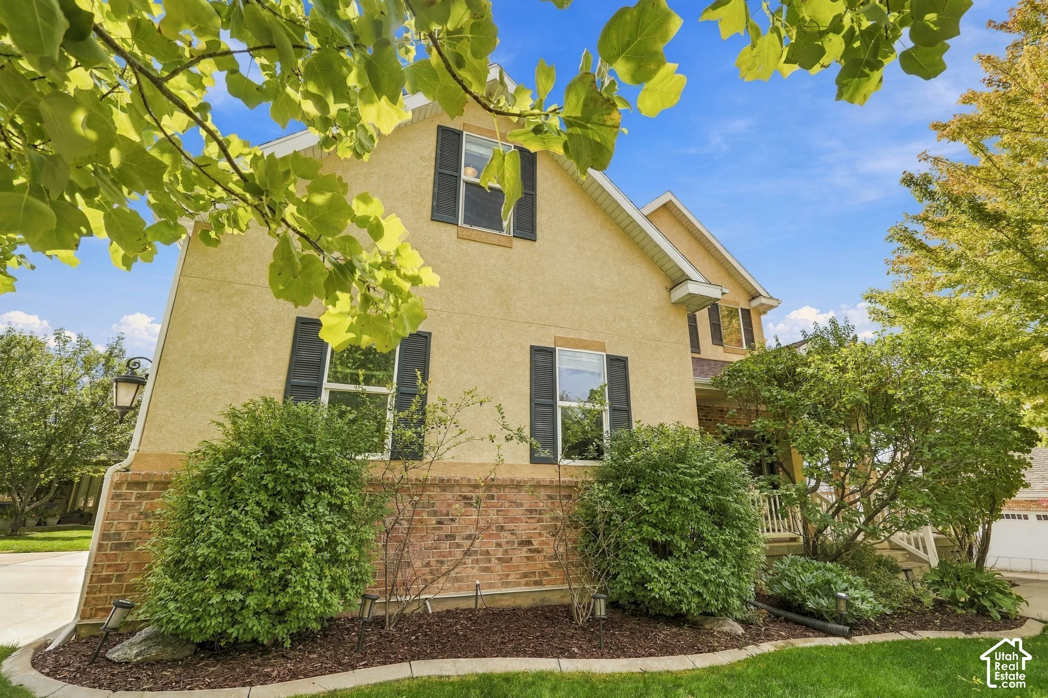 Stucco exterior with brick detailing, featuring multiple windows with black shutters, and mature landscaping