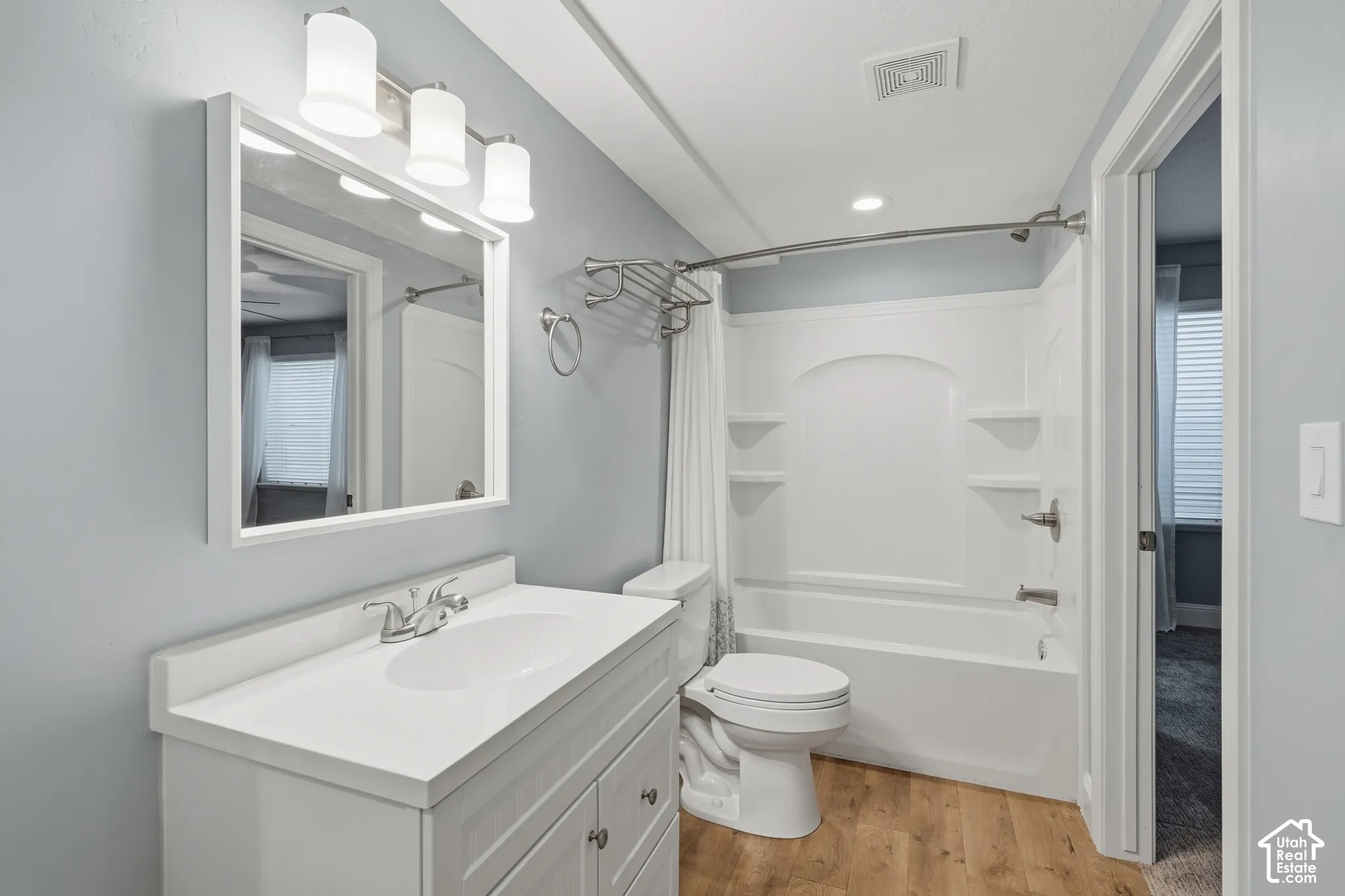 White vanity with integrated sink, wood-finish flooring, white framed mirror, three-light vanity fixture, and white tub/shower combination