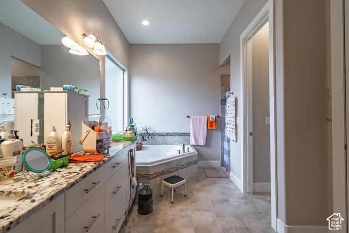 White vanity cabinetry with a light-toned countertop, a built-in corner tub with stone-finish surround, a large mirror, and tiled flooring