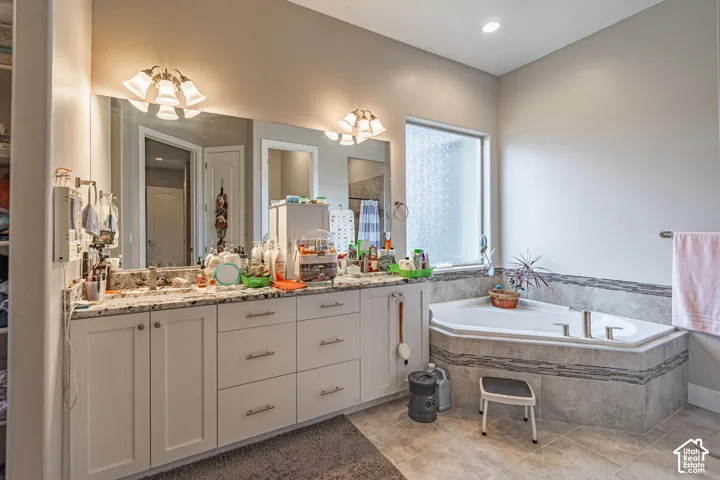 Dual vanity with stone countertop, large wall-mounted mirror, built-in corner tub, and textured tile flooring
