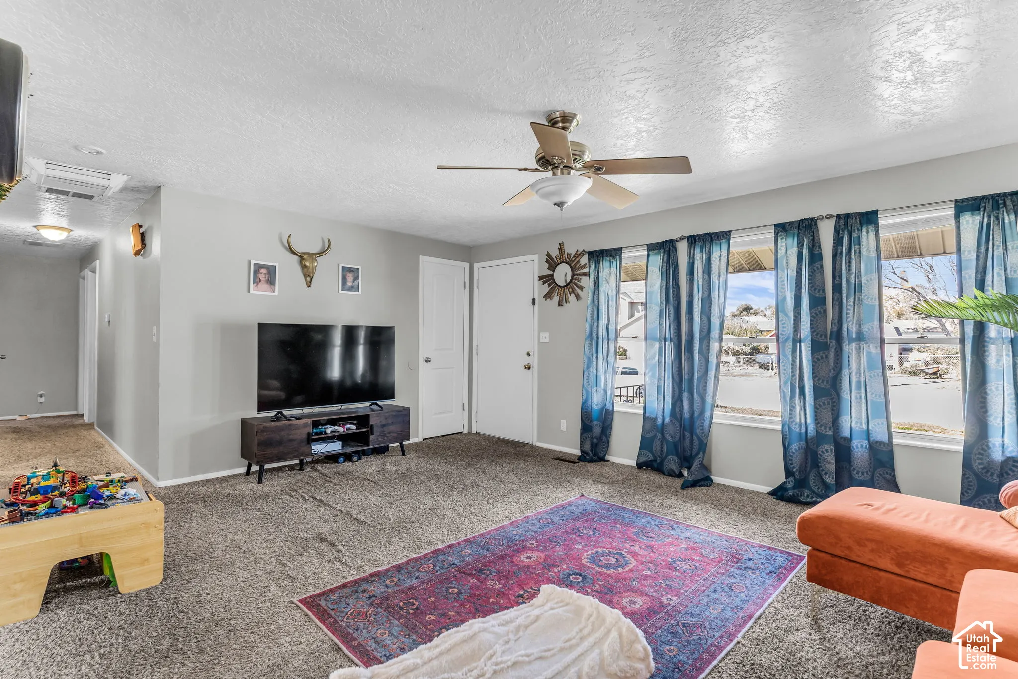 Spacious living area featuring neutral wall tones, carpeted flooring, a ceiling fan, and large windows