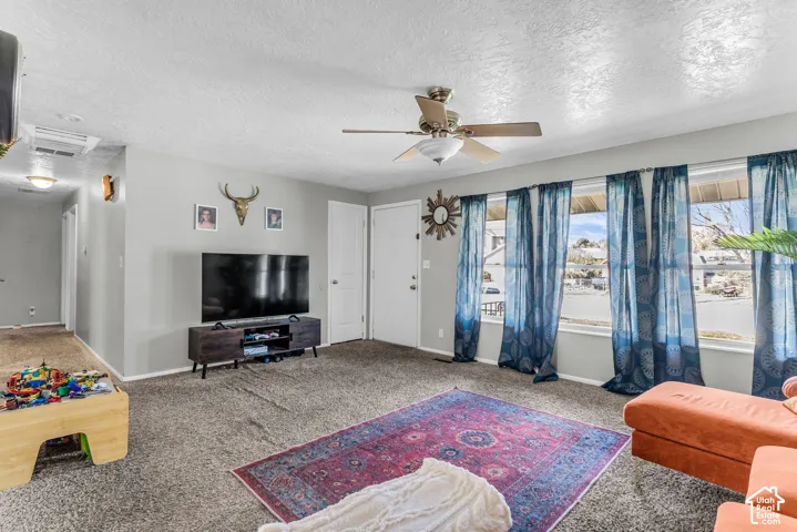 Spacious living area featuring neutral wall tones, carpeted flooring, a ceiling fan, and large windows
