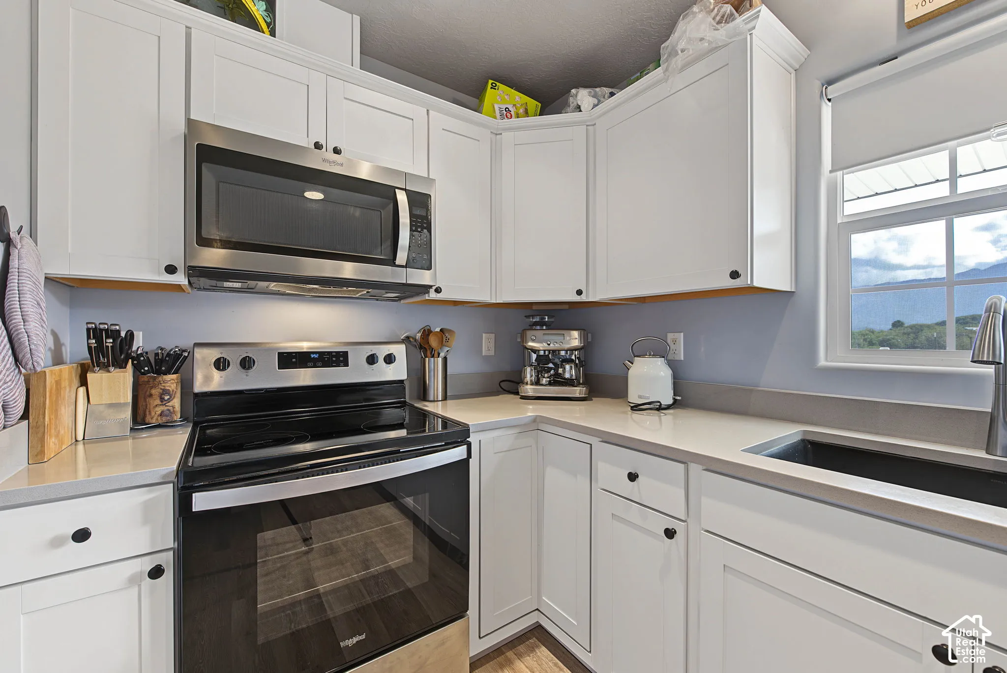 White shaker-style cabinetry with dark hardware, stainless steel range with overhead microwave, light-toned countertops, and an undermount sink with a gooseneck faucet