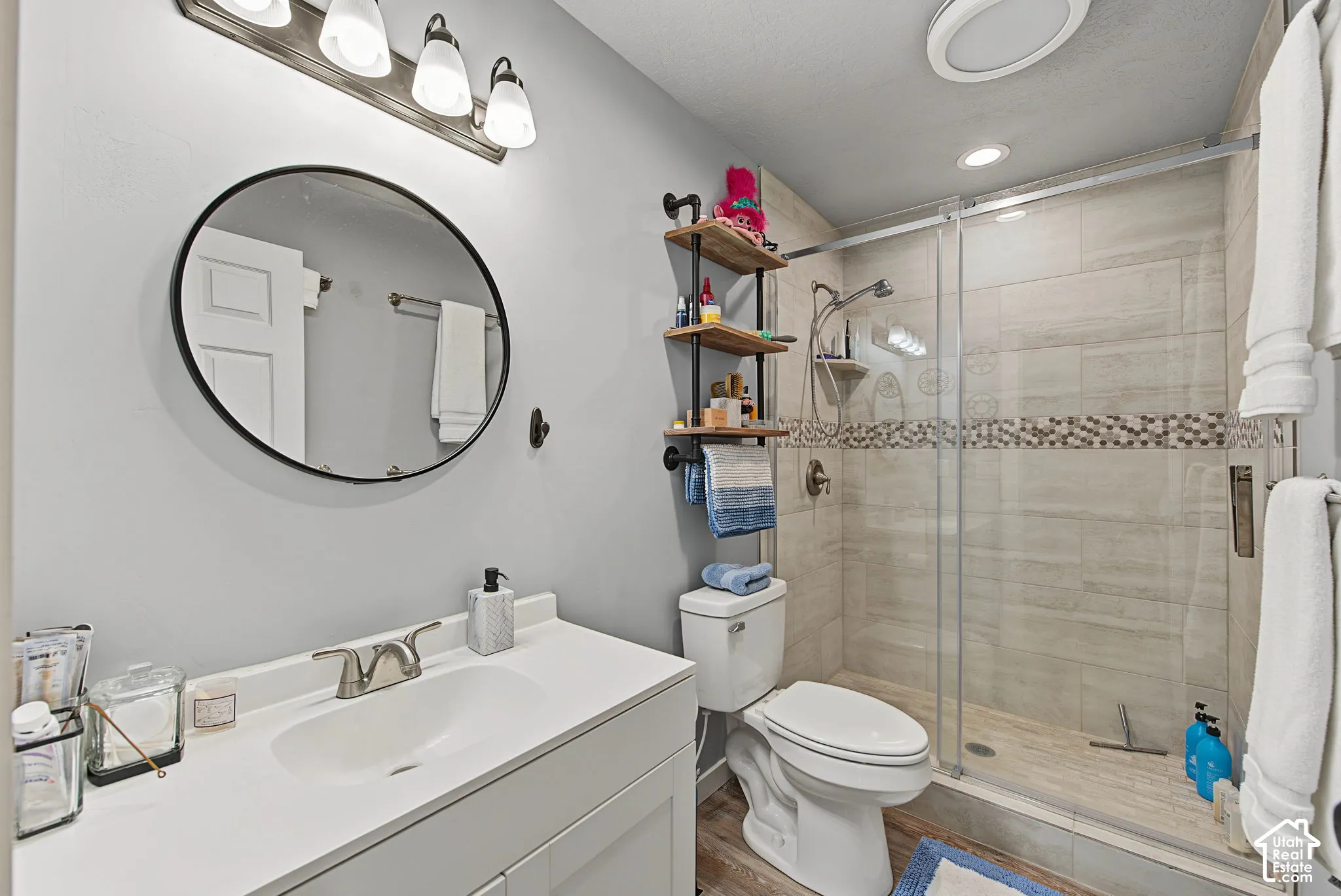 Bathroom featuring a single vanity with a white countertop, a circular framed mirror, a walk-in shower with glass enclosure, and tiled walls