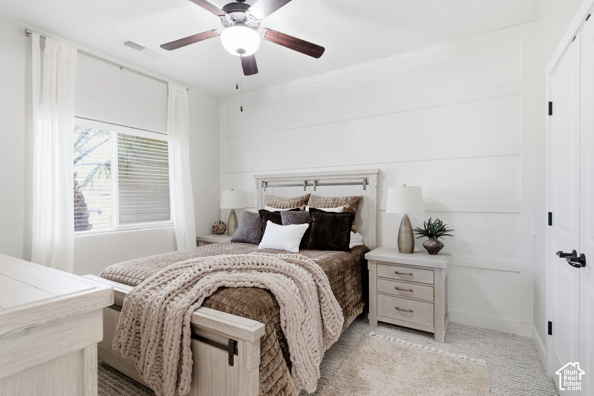 White paneled accent wall, neutral tone carpet, ceiling fan with integrated lighting, window with blinds, and white door with dark hardware