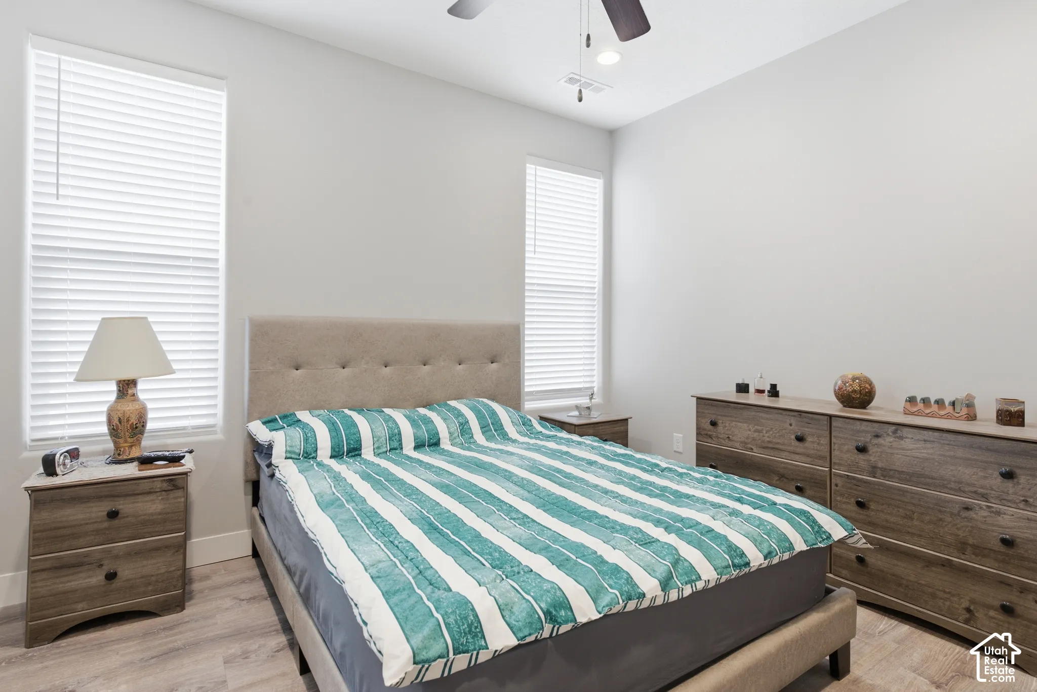 Neutral wall color, wood-finish flooring, ceiling fan with integrated lighting, two windows with blinds, and a tufted headboard