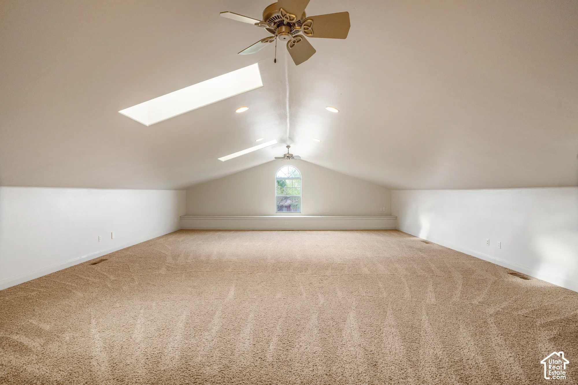 Expansive carpeted room featuring a vaulted ceiling, twin skylights, recessed lighting, ceiling fans, and an arched window