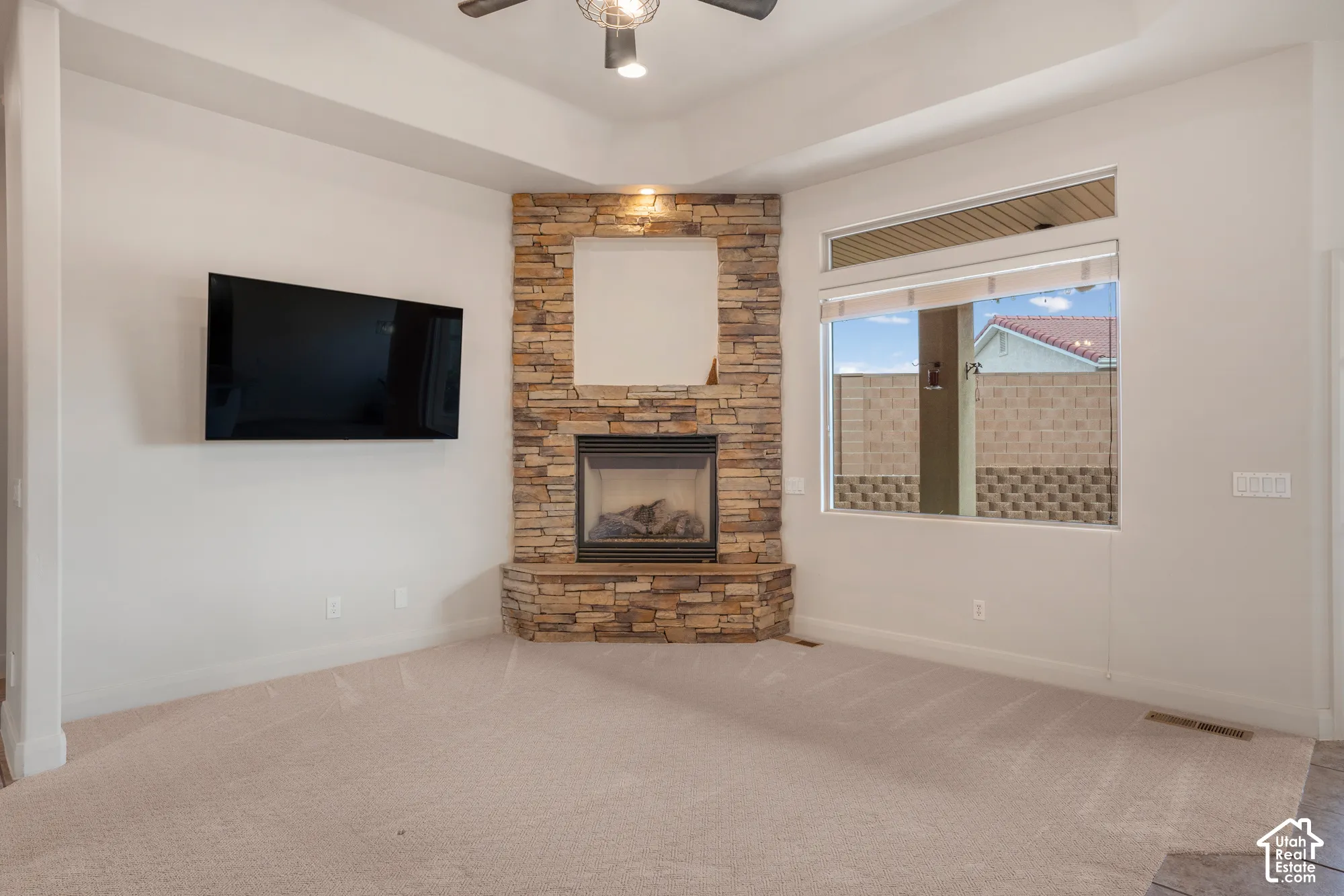 Corner stone fireplace with a raised hearth, large windows, tray ceiling, and ceiling fan with integrated lighting