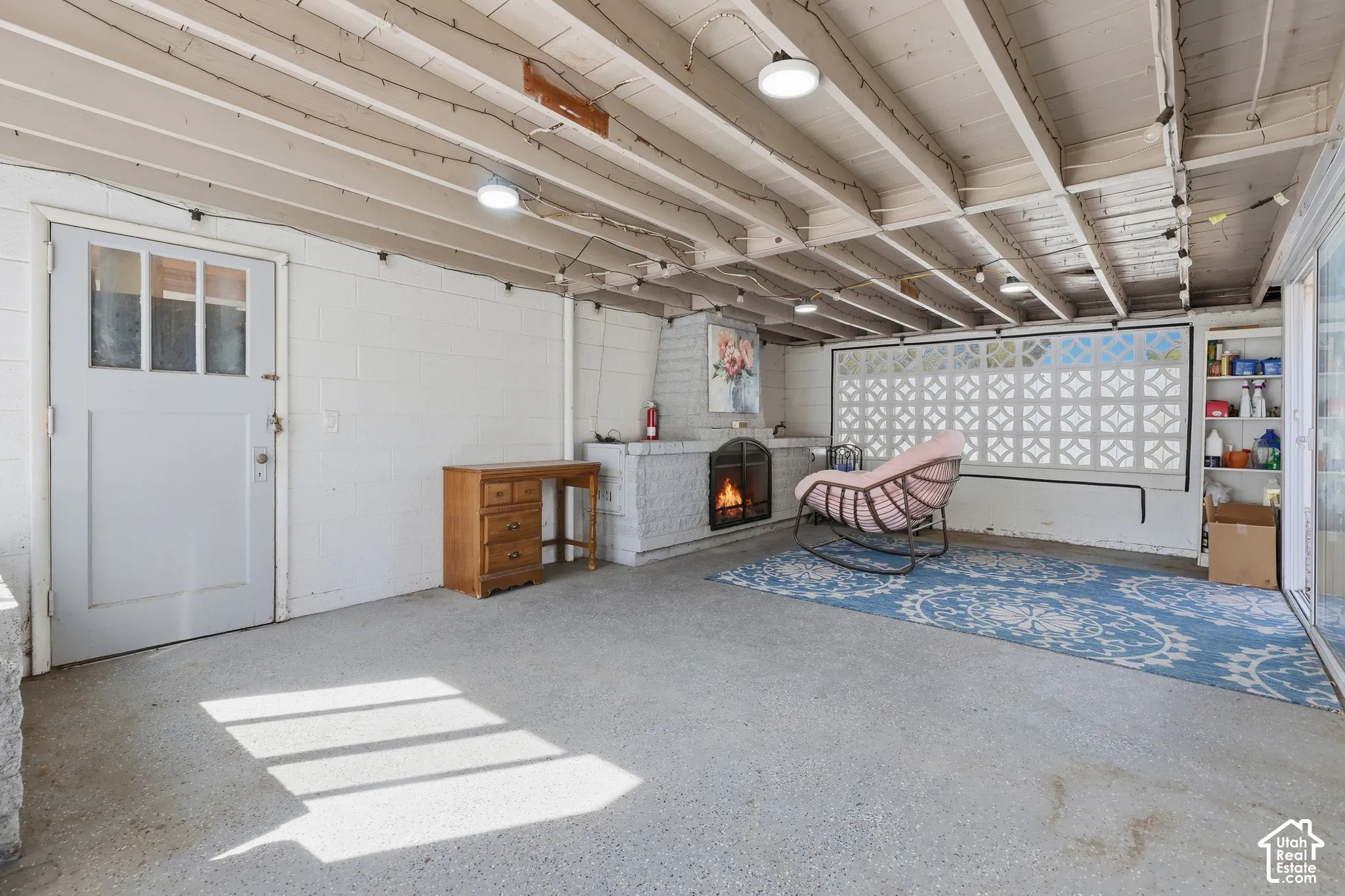 Exposed wood beam ceiling, white-painted brick fireplace, built-in shelving, glass-paneled door, and concrete flooring