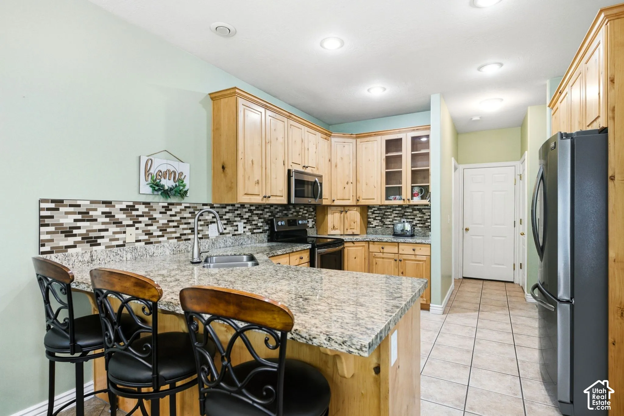 Kitchen featuring light wood cabinetry, a granite-finish island with seating, stainless steel sink, and a multi-tone tile backsplash