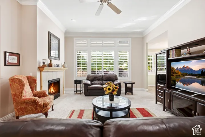 Carpeted living area featuring a tiled fireplace with mantel, crown molding, a ceiling fan, and plantation shutters