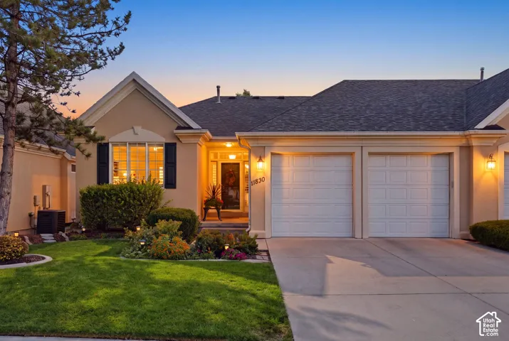 Stucco exterior with a dark shingle roof, offering a two-car garage, a concrete driveway, and established landscaping with accent lighting