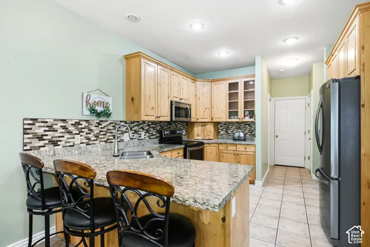 Kitchen featuring light wood cabinetry, a granite-finish island with seating, stainless steel sink, and a multi-tone tile backsplash