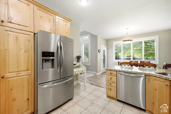 Full-size stainless steel refrigerator, light wood-finish cabinetry, light-toned tiled flooring, large windows with white louvered shutters, and a kitchen island with a light-toned countertop