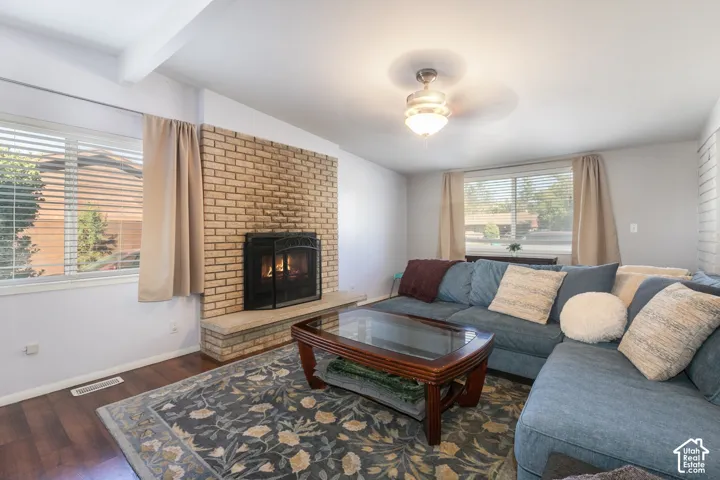 Brick-clad fireplace with hearth, wood-finish flooring, white ceiling beams, recessed lighting, and a ceiling fan with light fixture