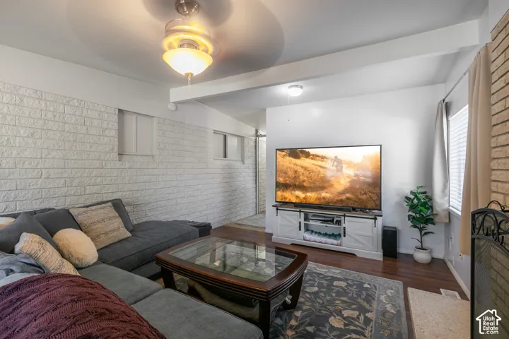 Recessed ceiling with an attached light fixture, a white painted brick accent wall, wood-finish flooring, and a window with blinds