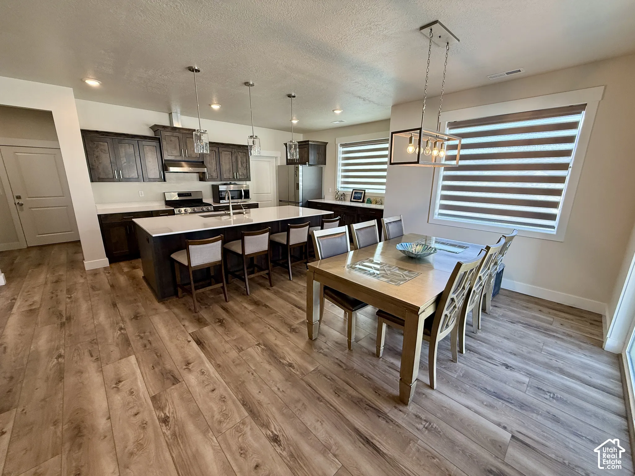 Open-concept living area with wood-finish flooring, a kitchen island with seating, dark wood cabinetry, stainless steel appliances, and multiple windows with blinds