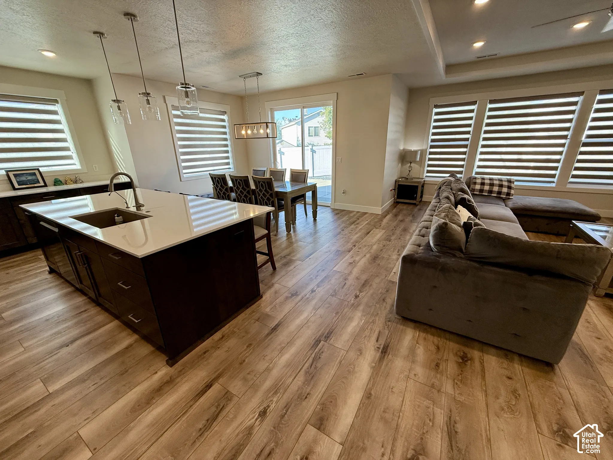 Open-concept living space with wood-finish flooring, a kitchen island with a sink and white countertop, recessed lighting, and large windows with horizontal blinds