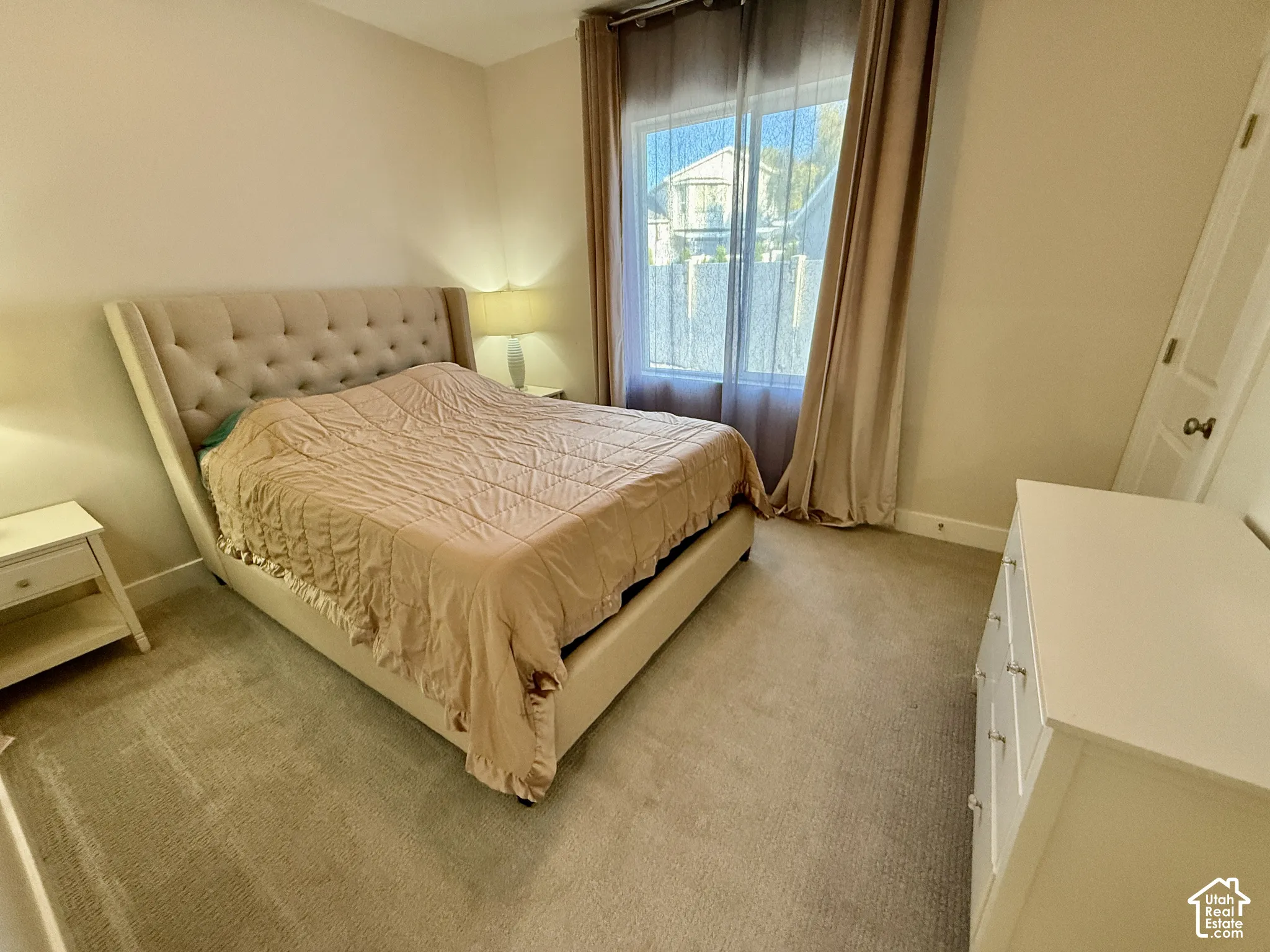 Neutral wall color, carpeted flooring, window with sheer and blackout drapes, white paneled door with round knob, white dresser with silver hardware