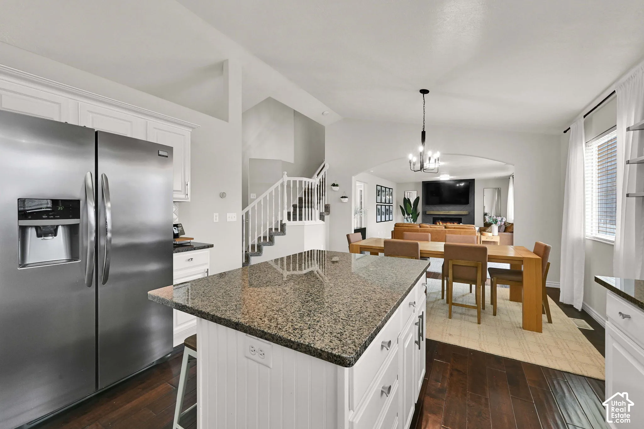 Kitchen island with granite countertop, stainless steel refrigerator, white cabinetry, dark wood flooring, and arch to family room